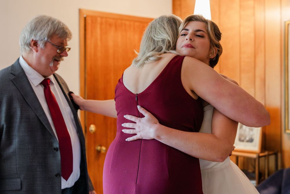 Three people share an emotional embrace during a wedding preparation moment in a wood-paneled room.