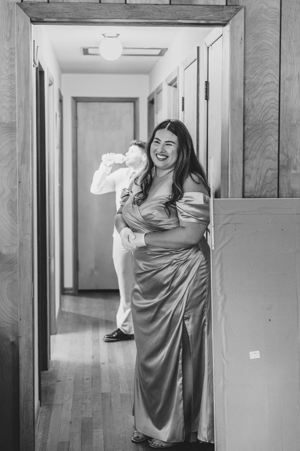 A person in an elegant long satin dress stands posed in a hallway, smiling joyfully during a special occasion.