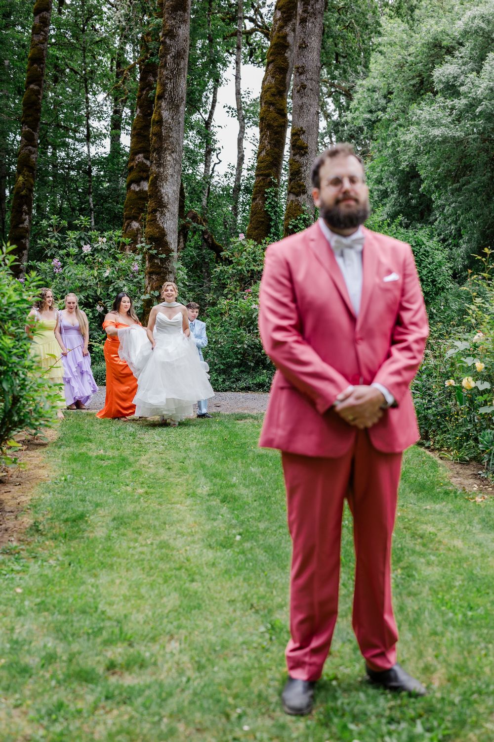 An outdoor wedding party in a forest setting with a bright coral pink suit and pastel bridesmaid dresses.