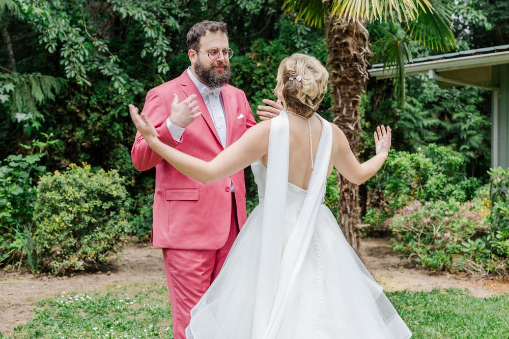 A couple dressed in a white gown and coral pink suit dance joyfully together in a lush garden setting.