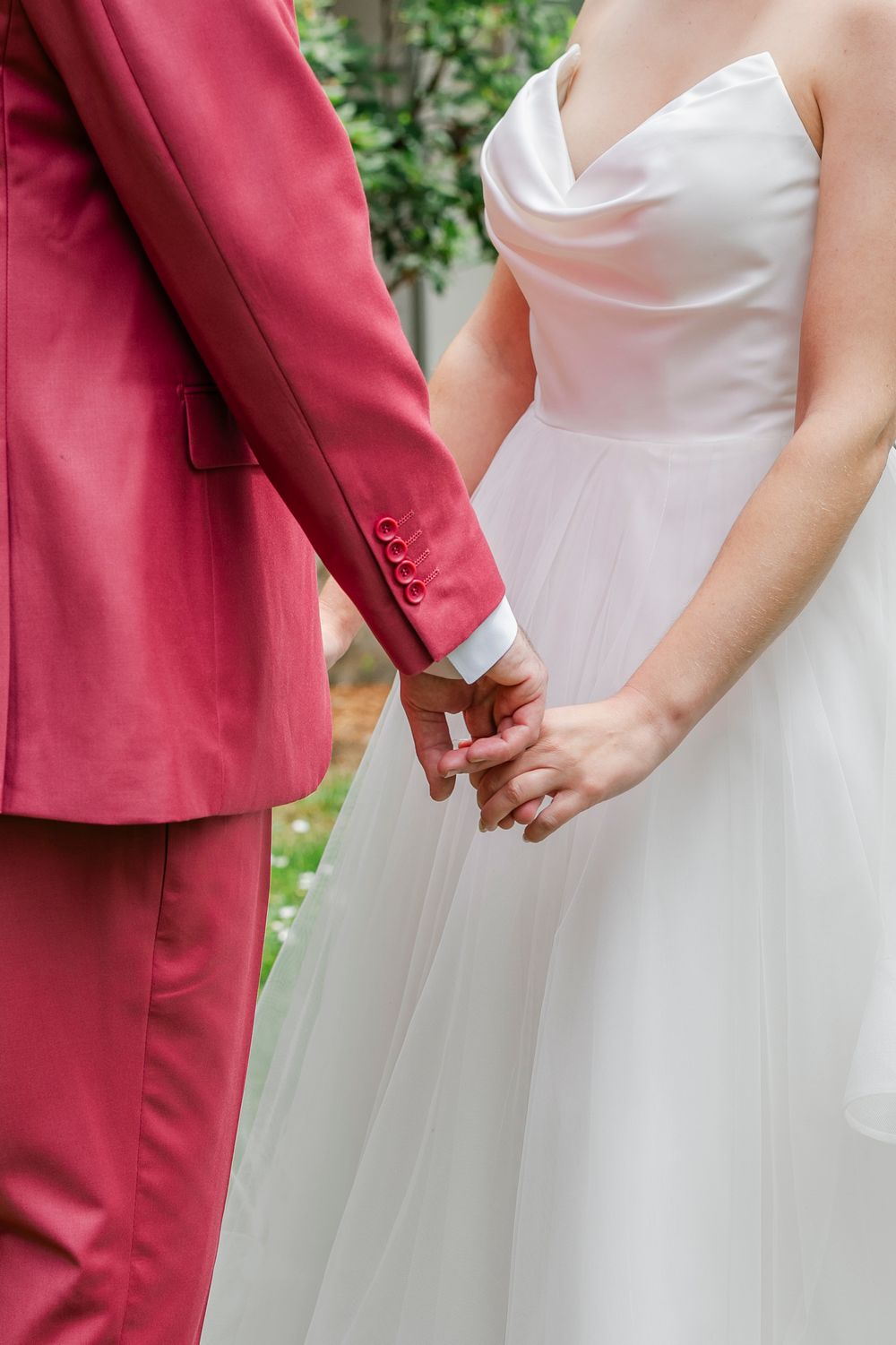 Close-up of couple holding hands, with bride in white tulle dress and groom wearing coral pink suit.