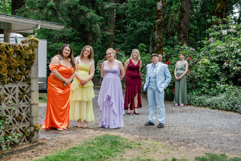 Bridal party wearing colorful dresses in orange, yellow, purple, burgundy and blue poses on gravel path by garden trellis.