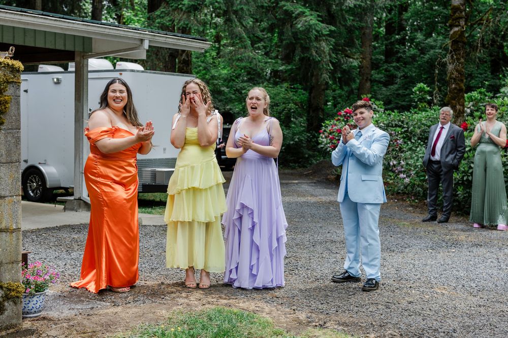 Wedding party members in colorful dresses and suits share an emotional moment on a gravel path near trees and a trailer.
