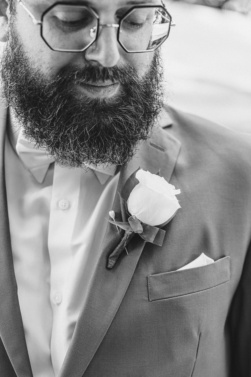 Close-up black and white photo of a bearded man wearing glasses and a white boutonniere.