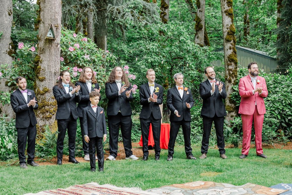 A wedding party in suits and a pink jacket pose together on grass with trees in the background.