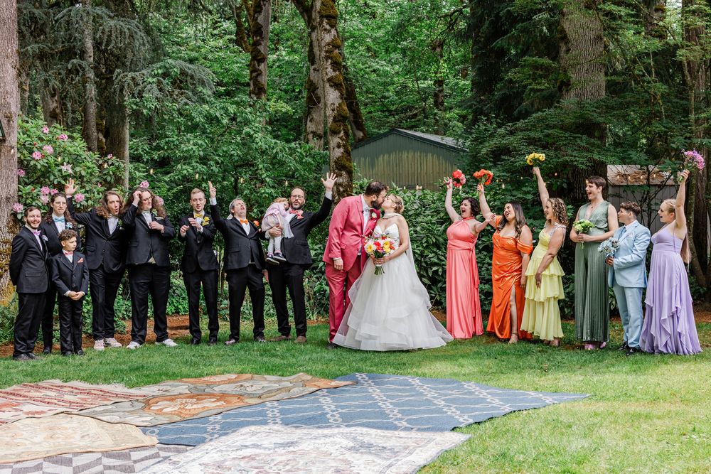 Wedding party in rainbow-colored dresses poses on stone patio surrounded by lush forest scenery.