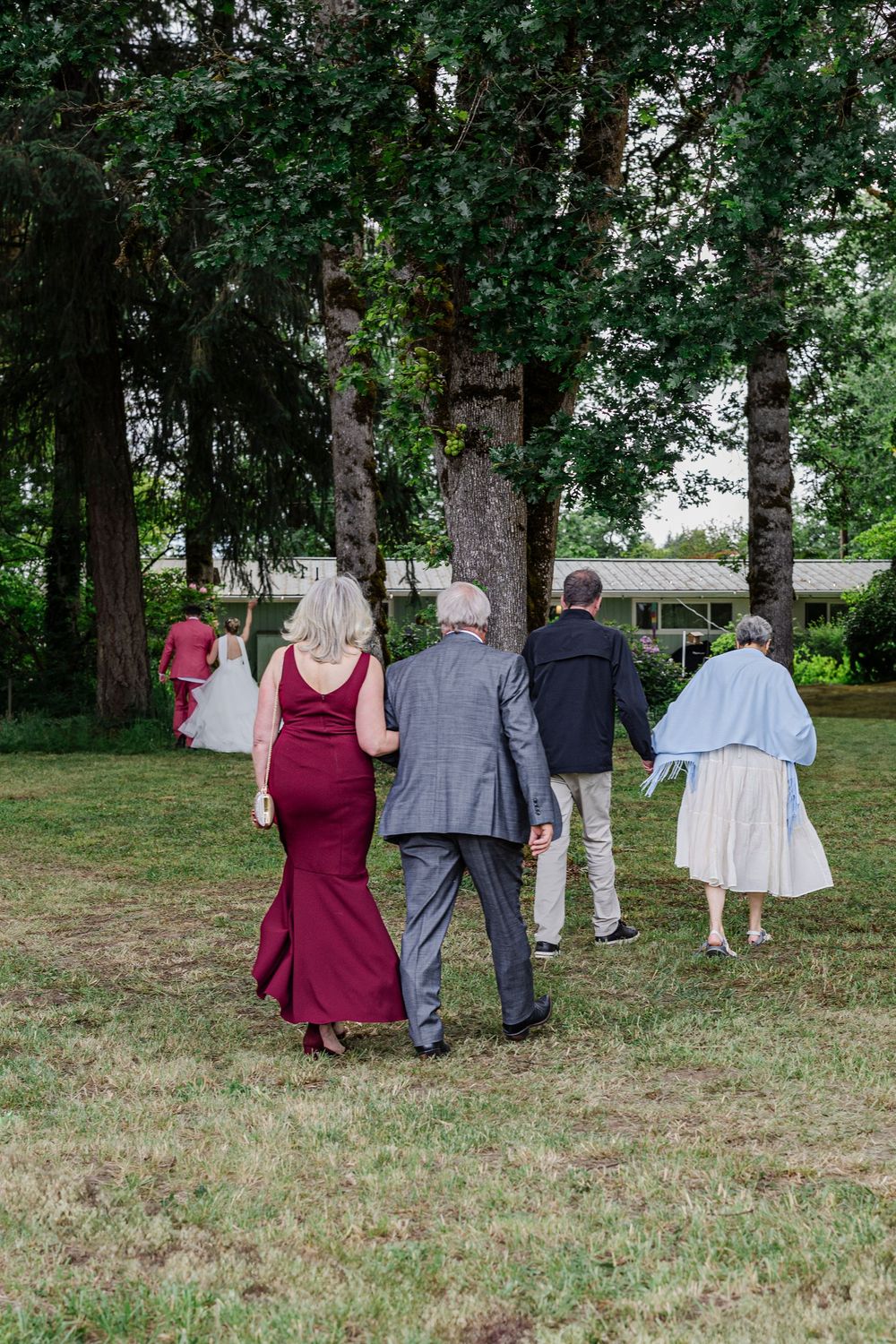 Wedding guests in formal attire walk across a grassy lawn surrounded by tall trees.