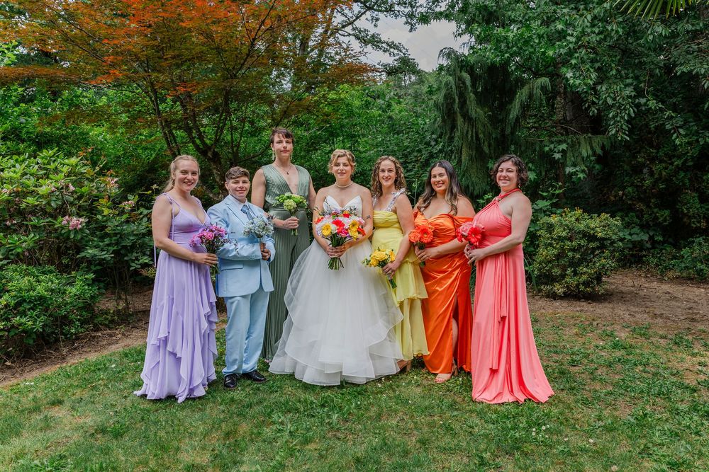 Bridesmaids wearing colorful dresses in shades of coral, yellow, purple and orange stand on grass in outdoor garden setting.