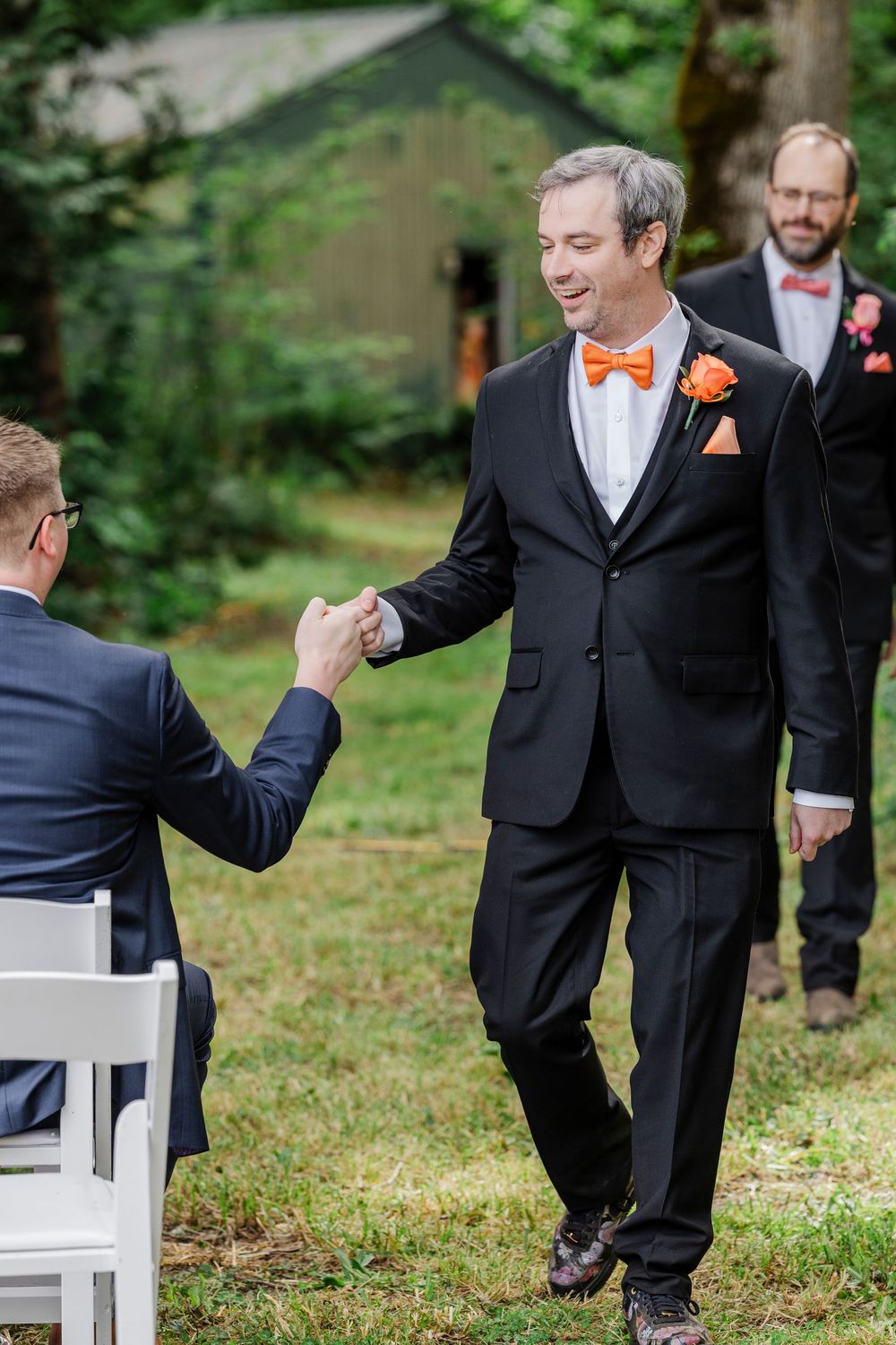 Members of a wedding party in black suits and orange bow ties engaged in a playful dance line on a grassy lawn.