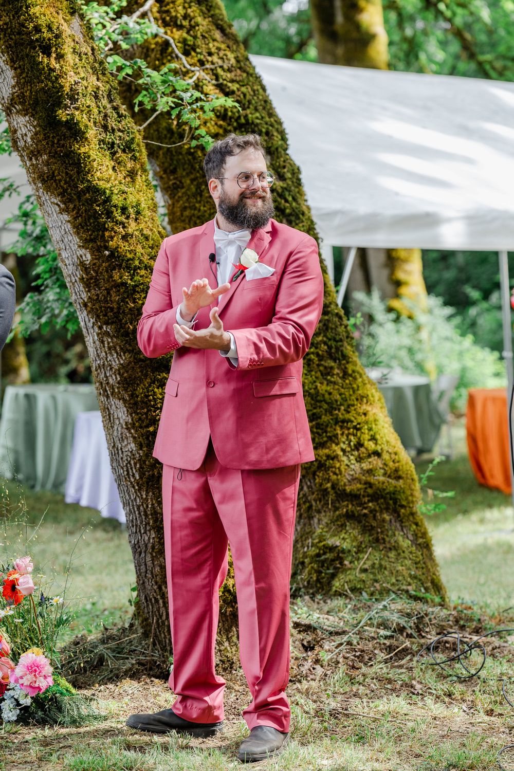 Wedding guest in bright coral suit stands beside moss-covered tree at outdoor ceremony.