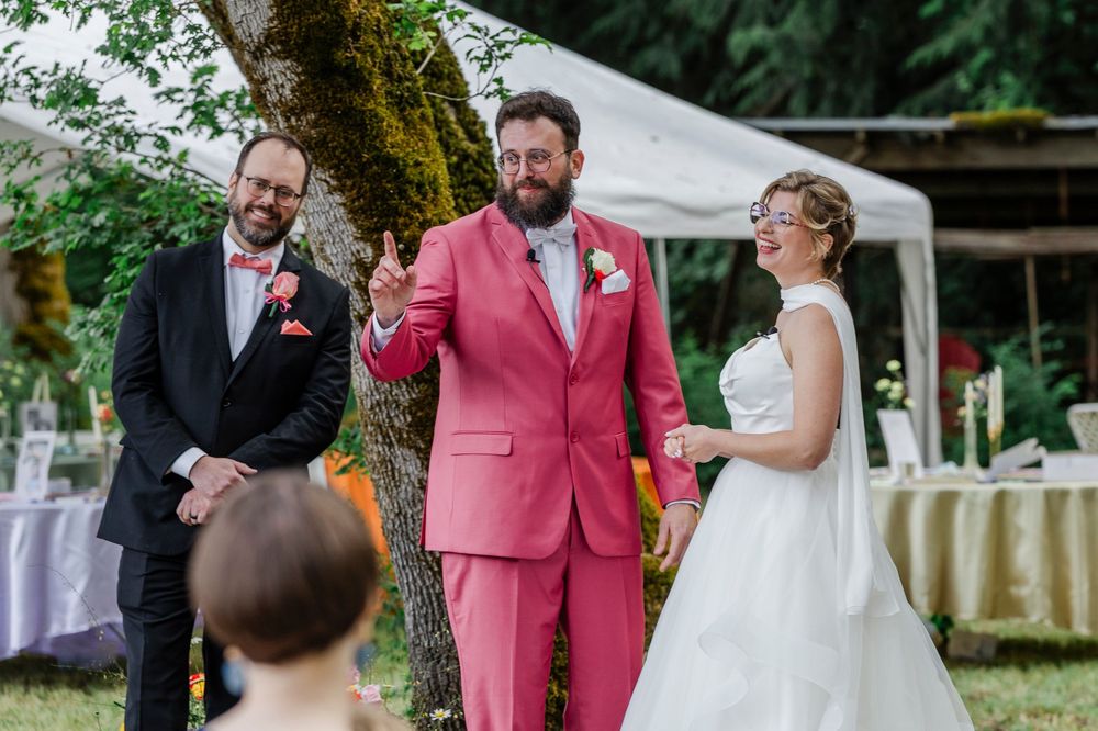 An outdoor wedding ceremony under trees with formal attire and a distinctive pink suit.