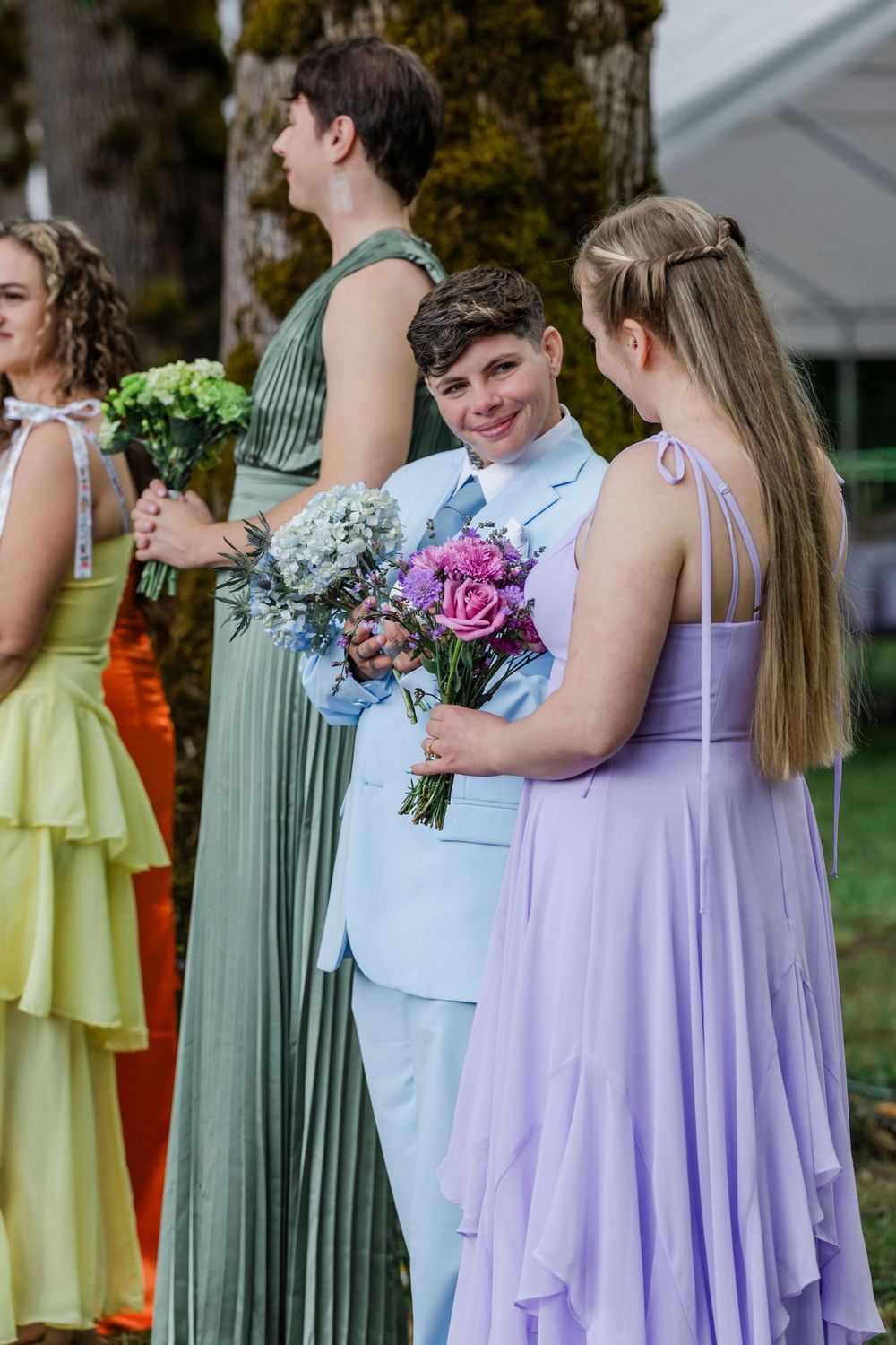 Wedding party members in pastel dresses and suits stand together holding colorful bouquets of flowers at outdoor ceremony.
