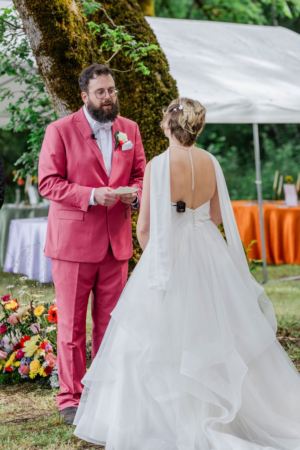 A couple at an outdoor wedding ceremony, with the groom in a pink suit reading vows under a white tent canopy.
