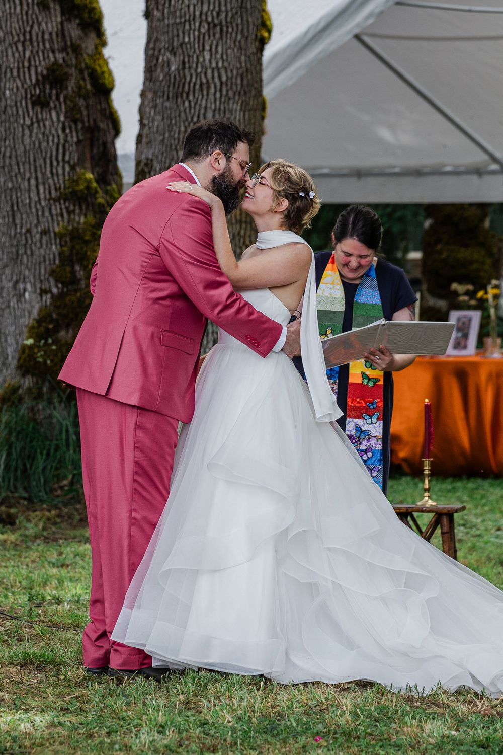 A couple in wedding attire share a romantic moment outdoors in front of trees while an officiant looks on.