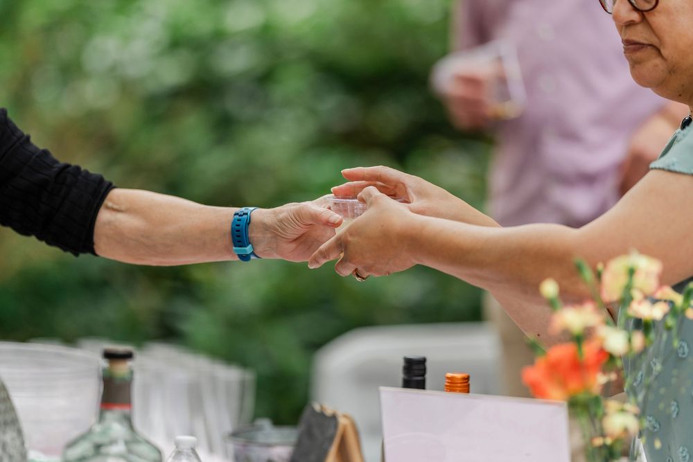 Close up of hands exchanging an item at an outdoor table with glasses and orange flowers in the background.