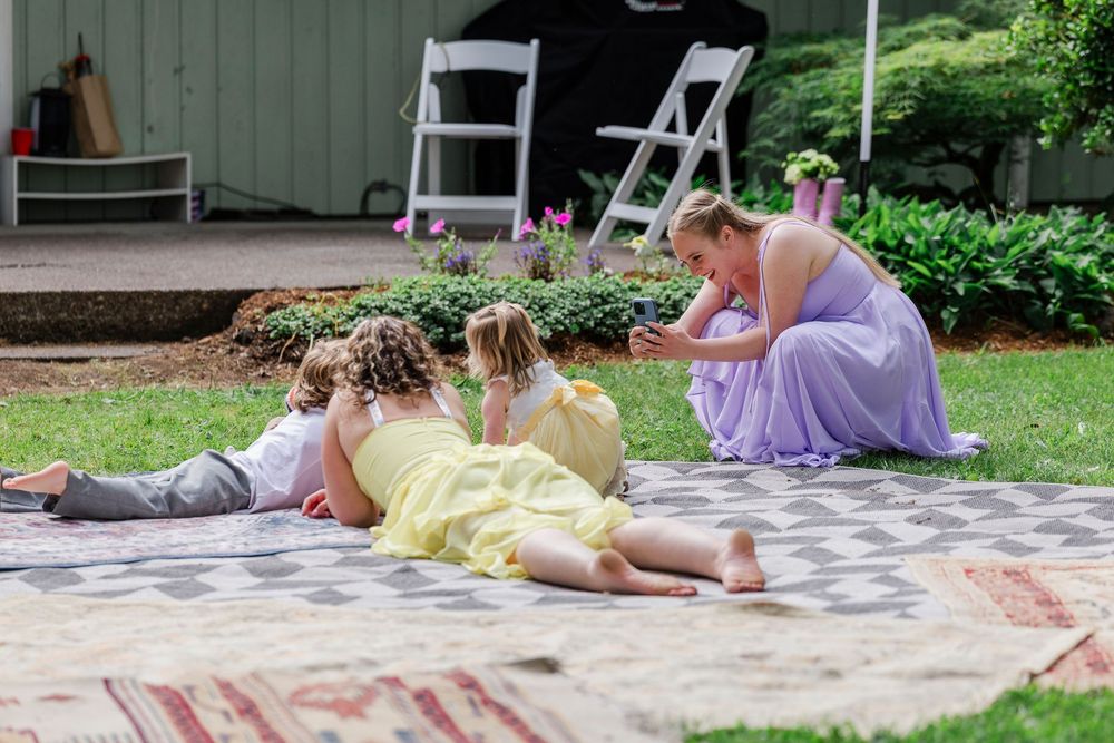 People in formal attire relax on a blanket at an outdoor garden gathering on a summer afternoon.