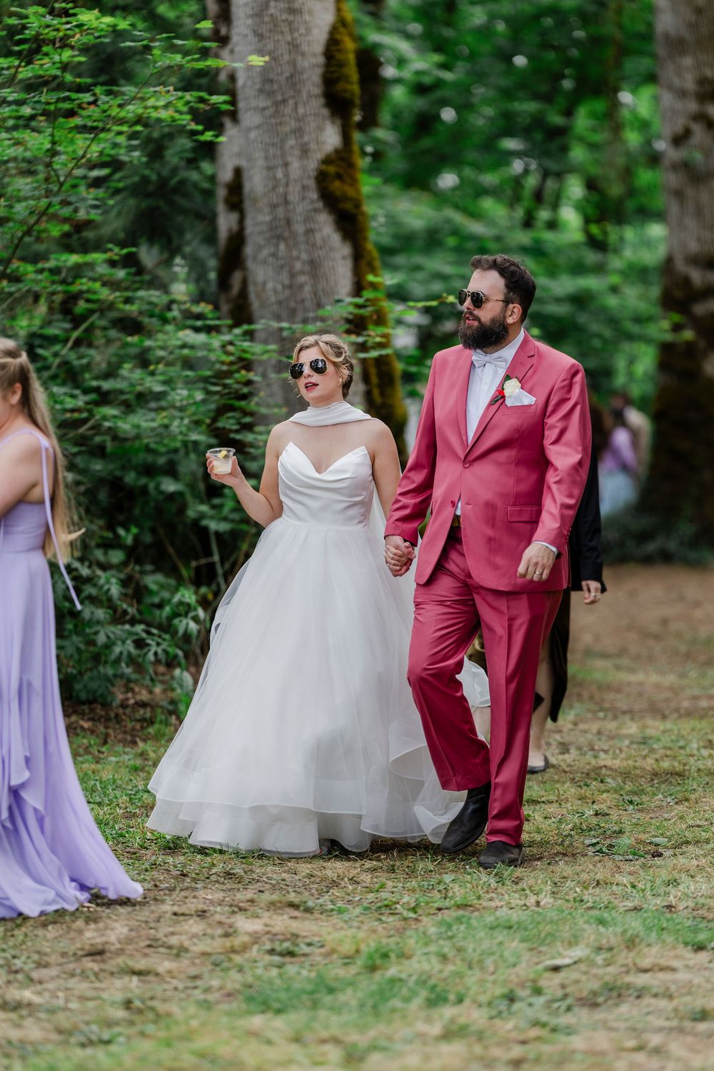 Wedding couple in rustic outdoor setting with bride in white gown and groom wearing stylish pink suit walking together.