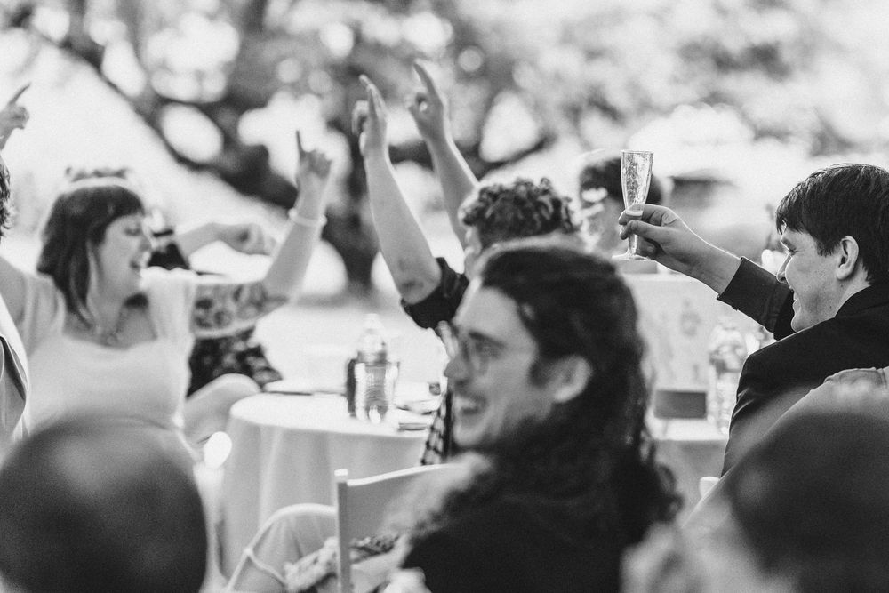 A black and white photo shows people celebrating at a wedding reception with raised arms and drinks at tables.