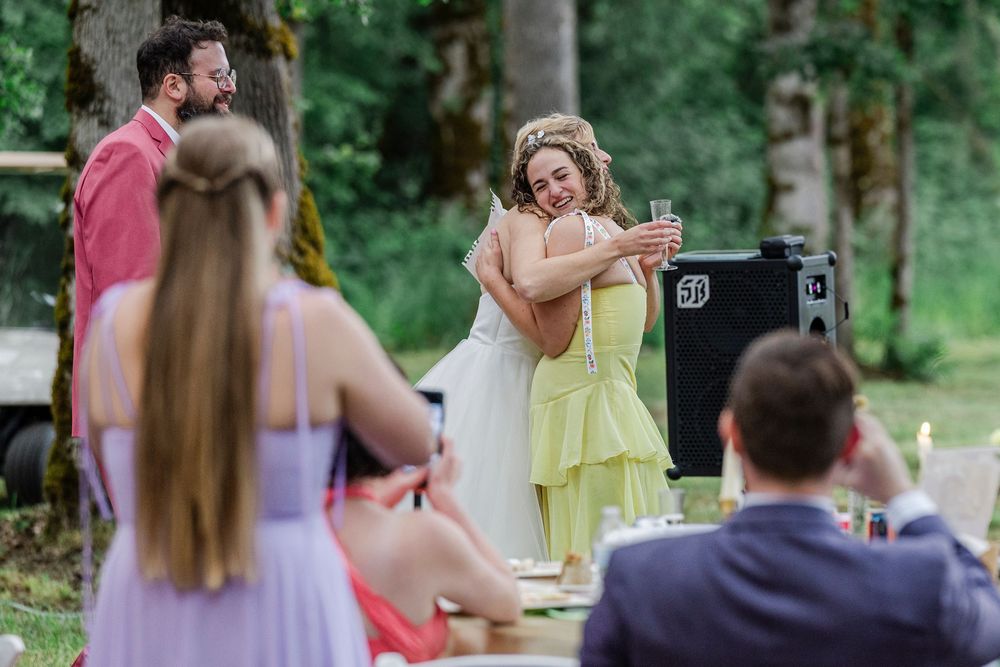 People gather for an outdoor celebration in a wooded setting with attendees in formal attire enjoying the festivities.