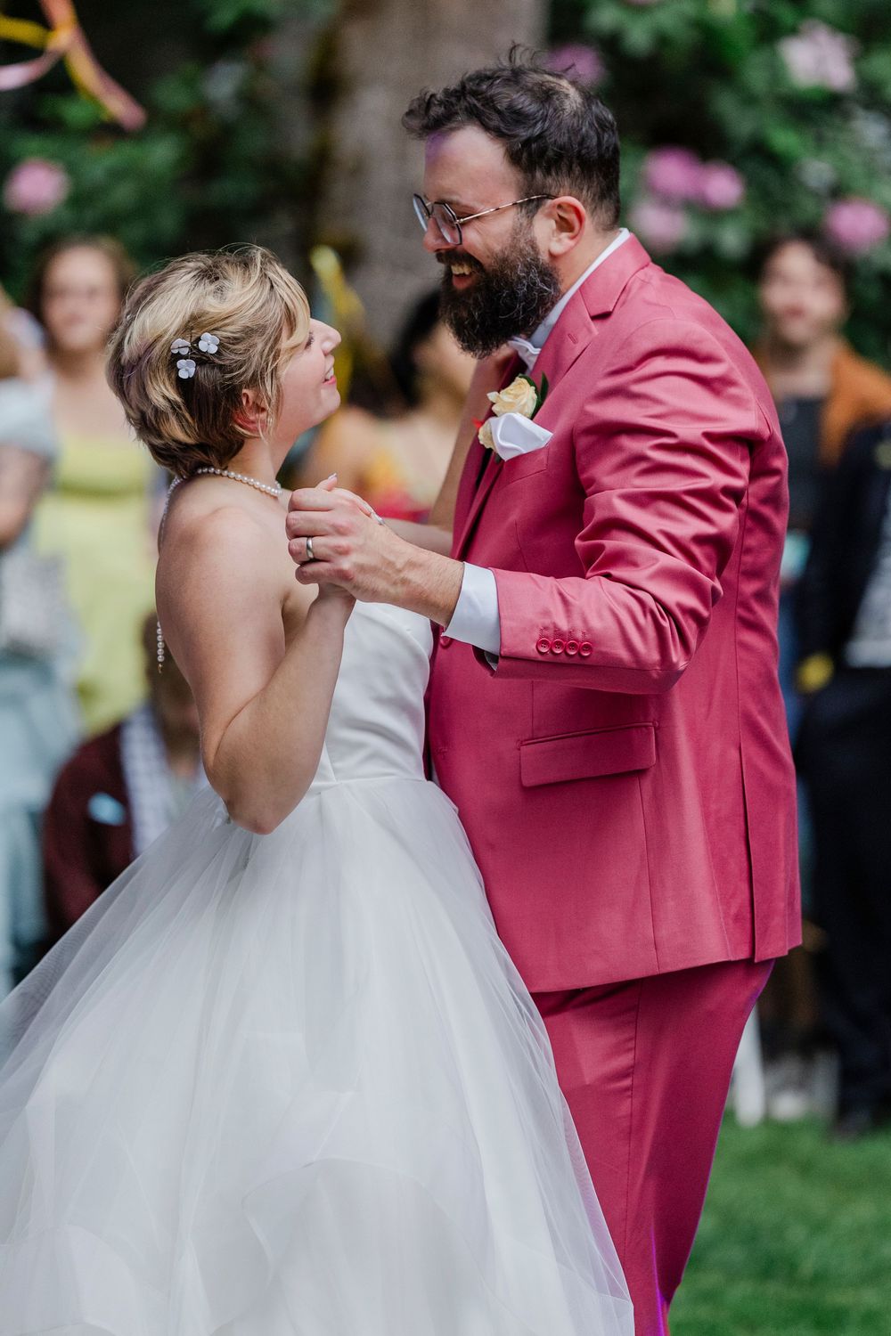A joyful couple share their first dance at an outdoor garden wedding, with the groom wearing a pink suit.