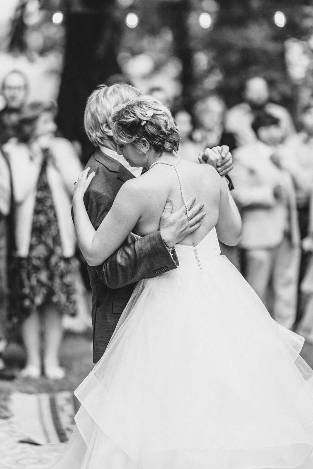 Black and white photo of newlyweds sharing their first dance at an outdoor wedding reception with guests watching.