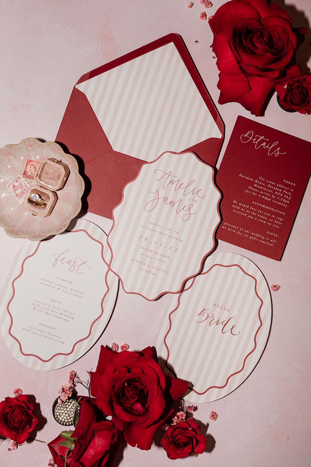 Elegant burgundy and white wedding invitation suite with scalloped edges and red roses against a pink background.