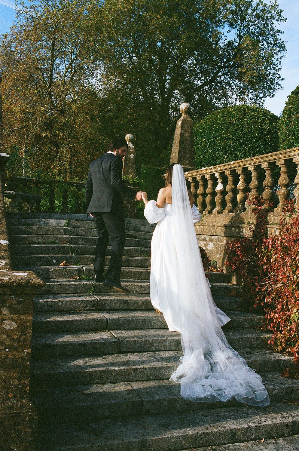 Two people in wedding attire descend stone garden stairs with lush greenery and ornate balustrade in romantic outdoor setting.