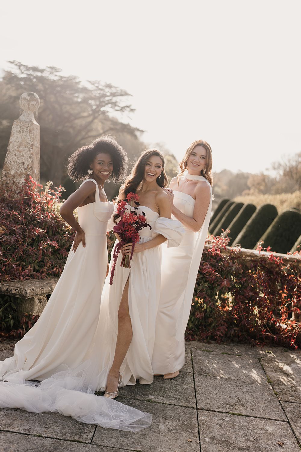 A group in white wedding dresses stands in a garden setting during golden hour with red bouquets.