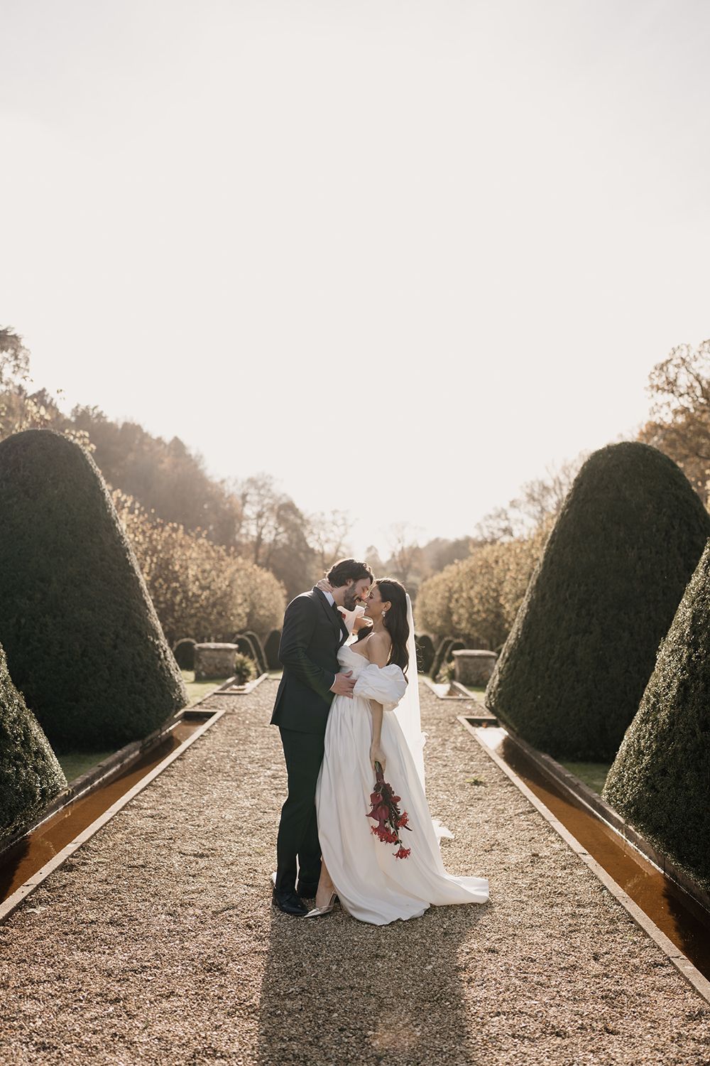 Newlyweds share an intimate moment on a garden path lined with manicured topiary hedges at sunset.