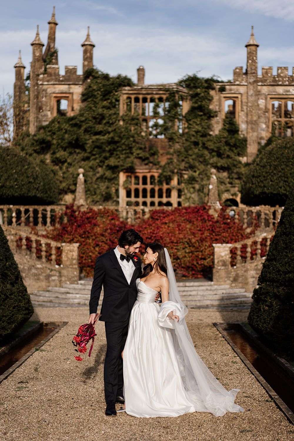 Newly married couple poses on castle grounds with stone walls and red autumn foliage in the background.