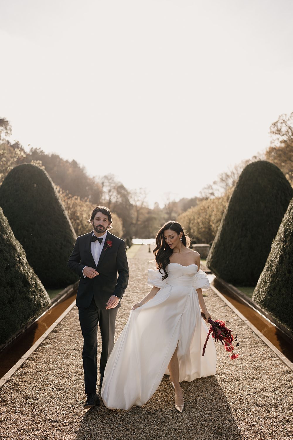 A couple shares a romantic moment while walking along a garden path lined with manicured hedges during a wedding photo session.