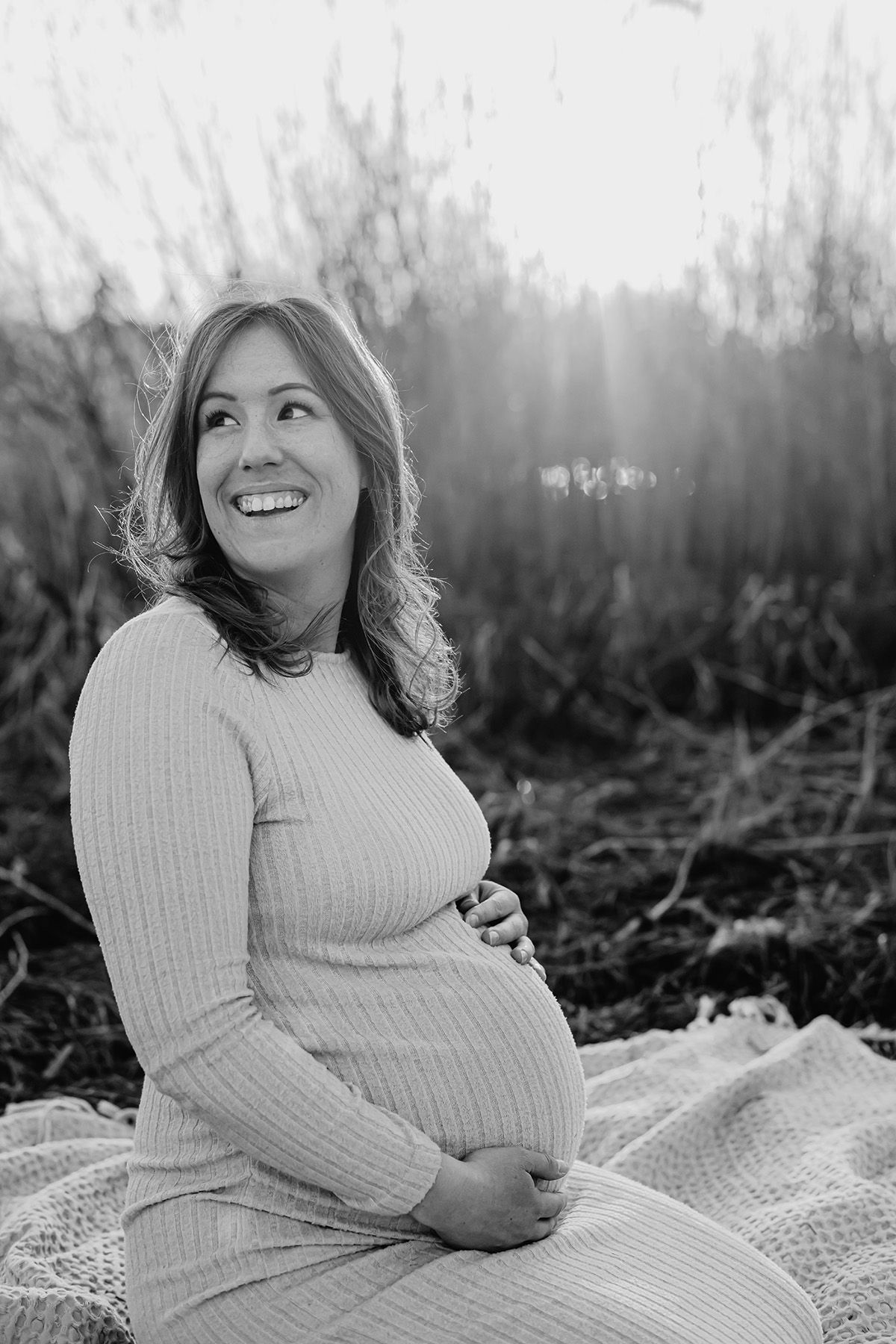 En gravid kvinna fotograferad utomhus sittand på en filt vid strandkanten, hon ler och håller en hand på magen, fotograf Josefin Landhagen.