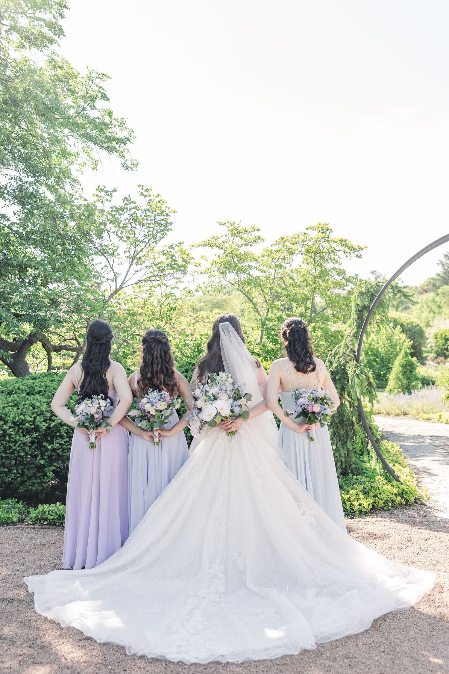 A bride in a white gown with long train stands with three bridesmaids in lavender dresses holding bouquets in a garden setting.