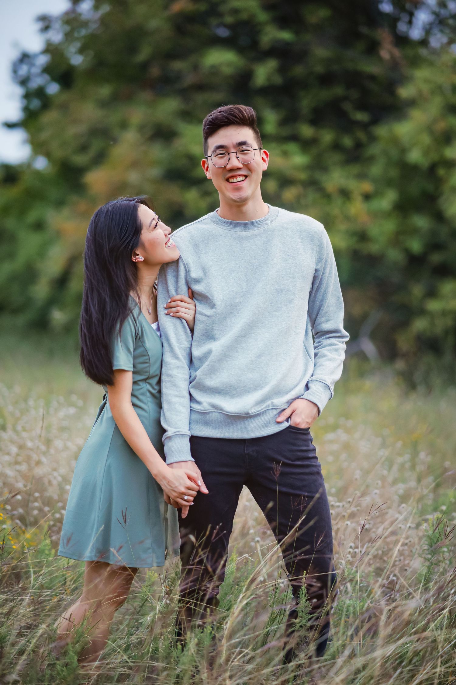 A romantic couple stands together in tall grass and wildflowers, wearing casual clothing on a sunny afternoon for engagement photos