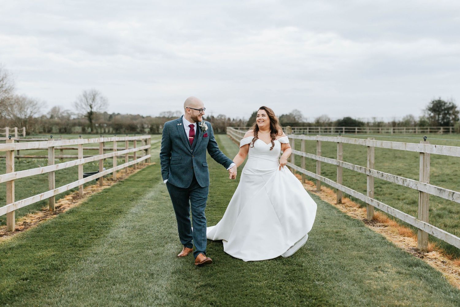 A couple walks hand in hand along a grassy path between white fences at an outdoor countryside wedding venue.