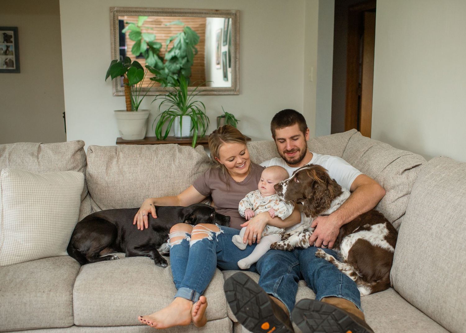 A family relaxes together on a light gray couch while playing and cuddling with their small brown dog.