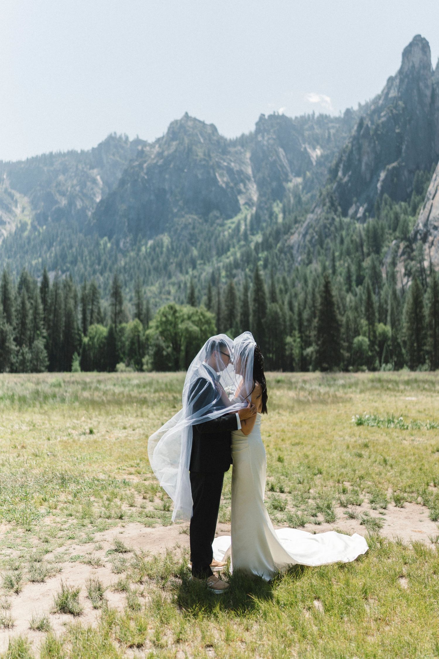 A wedding couple embraces under a flowing veil in a meadow with dramatic mountain peaks and evergreen forest behind them at yosemite valley