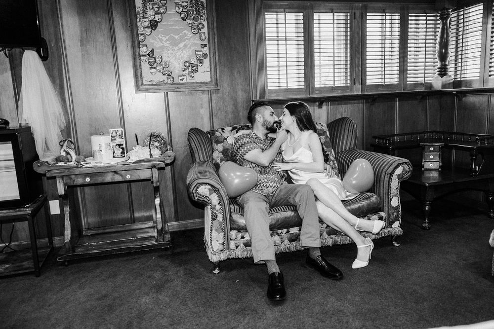 A couple in vintage 1950s clothing relaxes together on a floral armchair in a wood-paneled room with period decor.