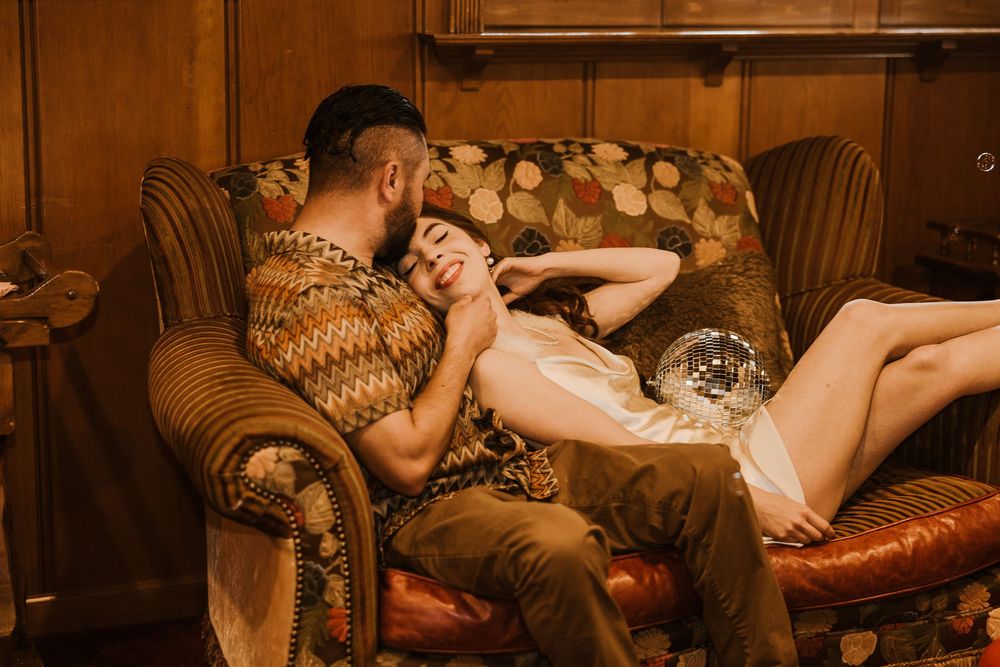 Couple lounges intimately on brown leather sofa with vintage decor in warm toned romantic setting.