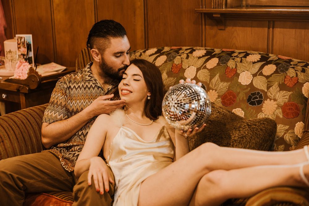 A romantic couple lounges together on a retro floral couch with a disco ball in warm sepia tones.