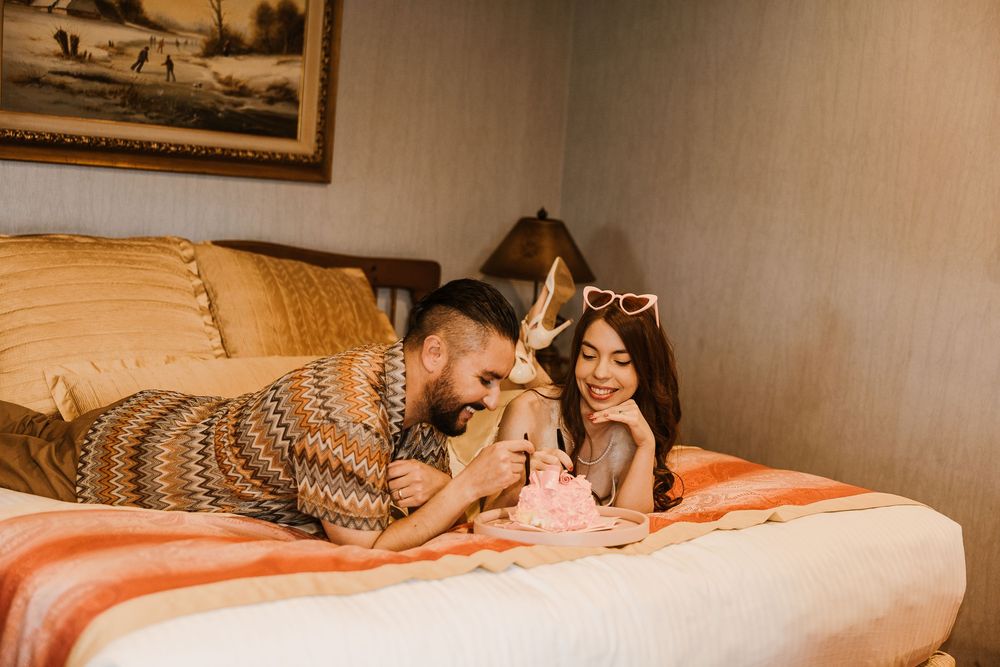 Two people lie on a bed smiling while looking at their phones in a cozy room with warm lighting.