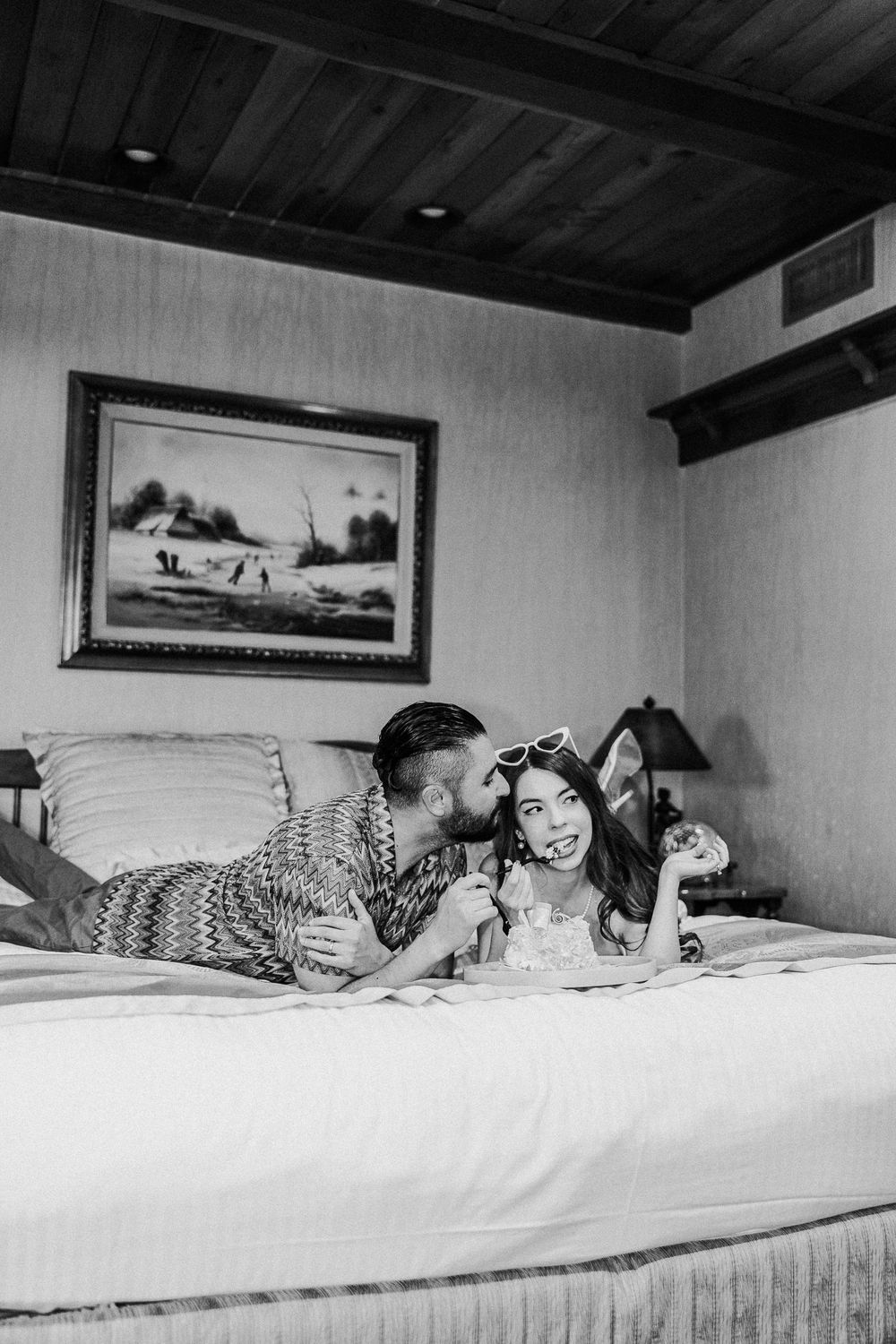 A loving couple shares a joyful intimate moment laying together on a white bed beneath a framed landscape artwork.