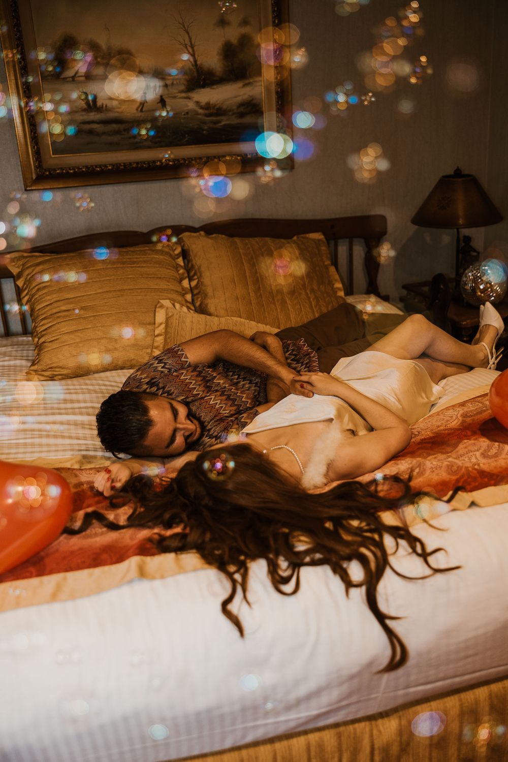 Person lying on bed with long curly hair against brown pillows in warm ambient lighting with bokeh effects.