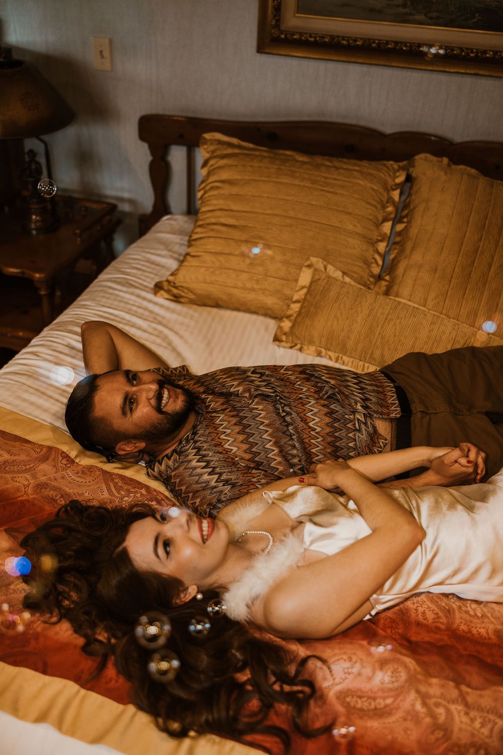 A couple rests on a bed with gold pillows and striped bedding in a cozy bedroom setting with warm lighting.
