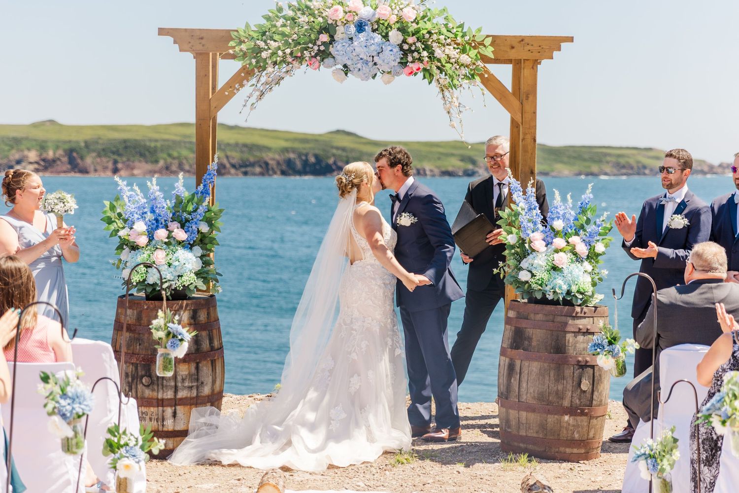 Rustic outdoor wedding ceremony by the ocean with wooden barrels and floral arch decorated in blue and white flowers.