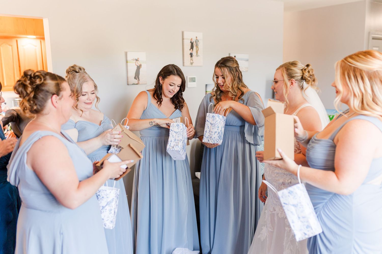 Several women in matching dusty blue bridesmaid dresses getting ready in a sunlit room together before a wedding ceremony.