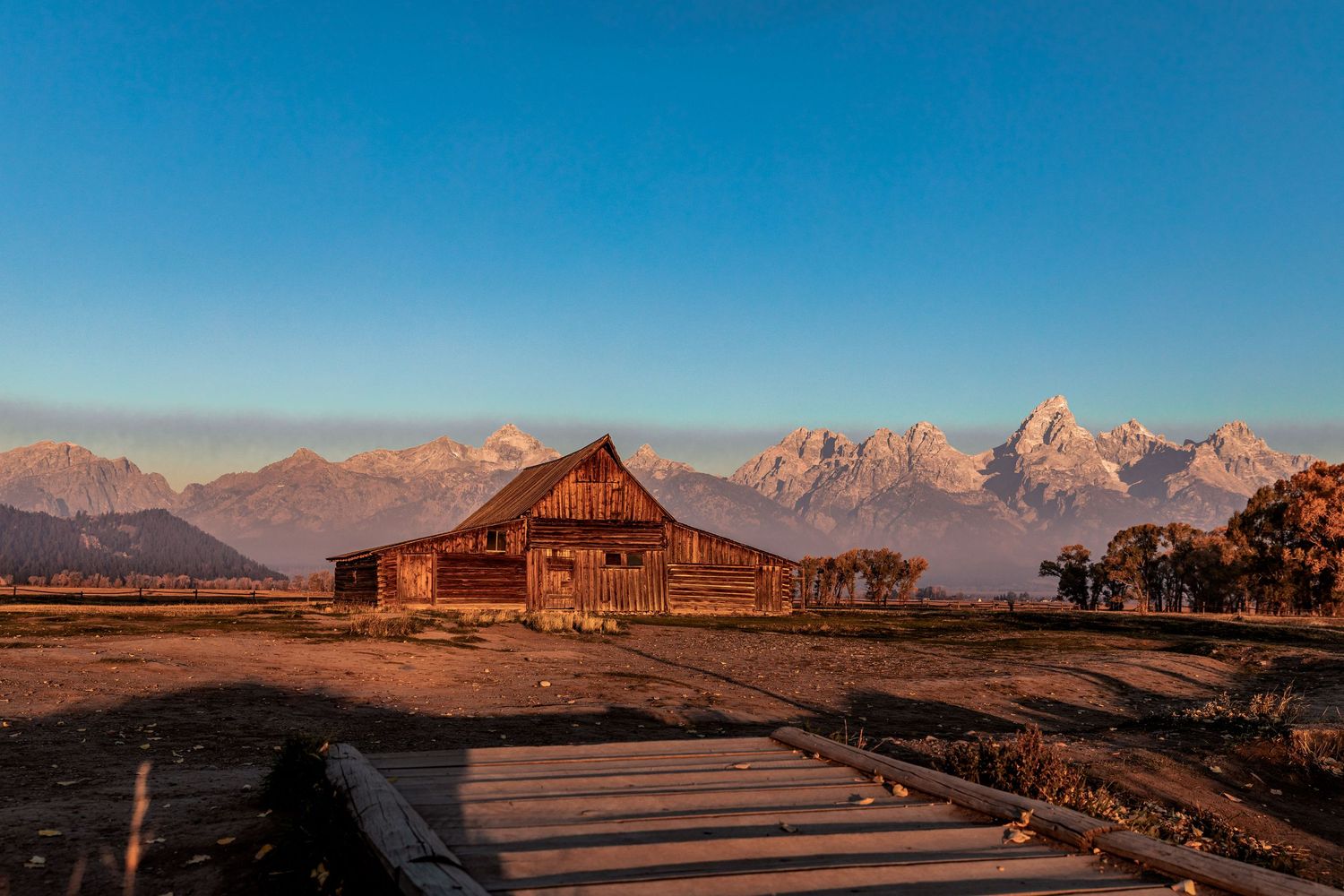 Rustic wooden barn stands against majestic Teton mountain range at sunset in Grand Teton National Park.