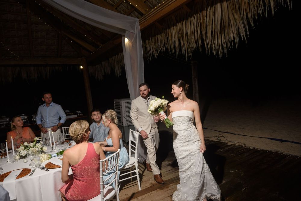 Wedding guests dance and celebrate at a nighttime tiki bar reception under thatched roof lighting.