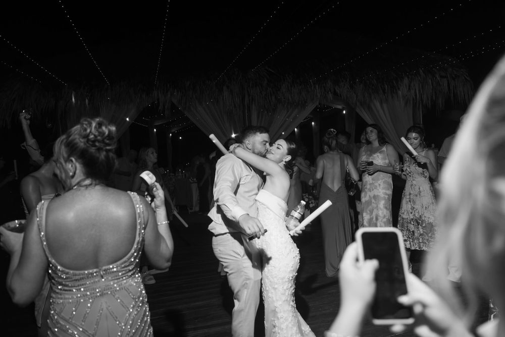 A couple in matching outfits dance together while guests surround them taking photos at a nighttime celebration.