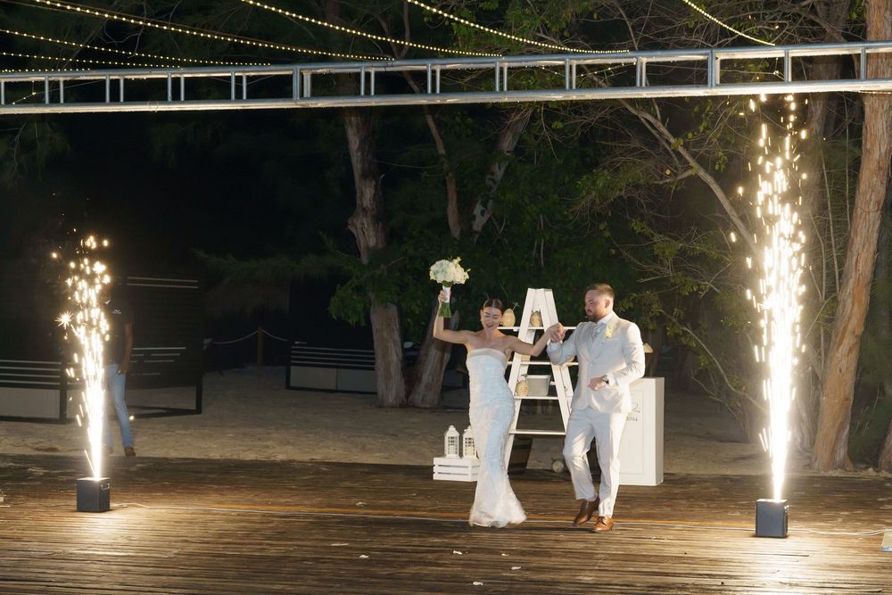Sparklers illuminate an elegant nighttime beachside wedding ceremony setup with white decor on a wooden deck.